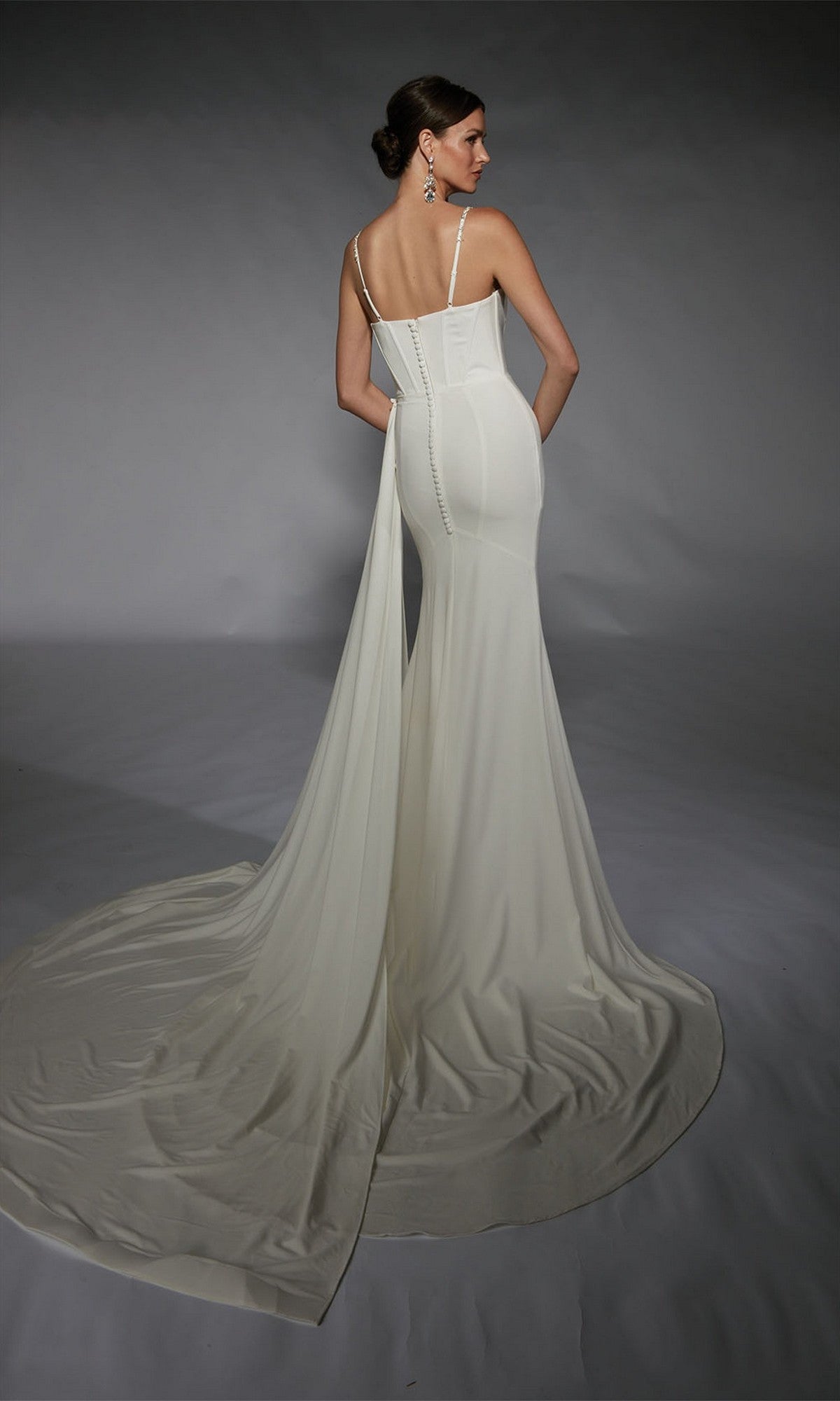 A woman is seen from behind wearing the Alyce 7104, a fitted floor-length white sheath dress with a long train, standing against a gray background.